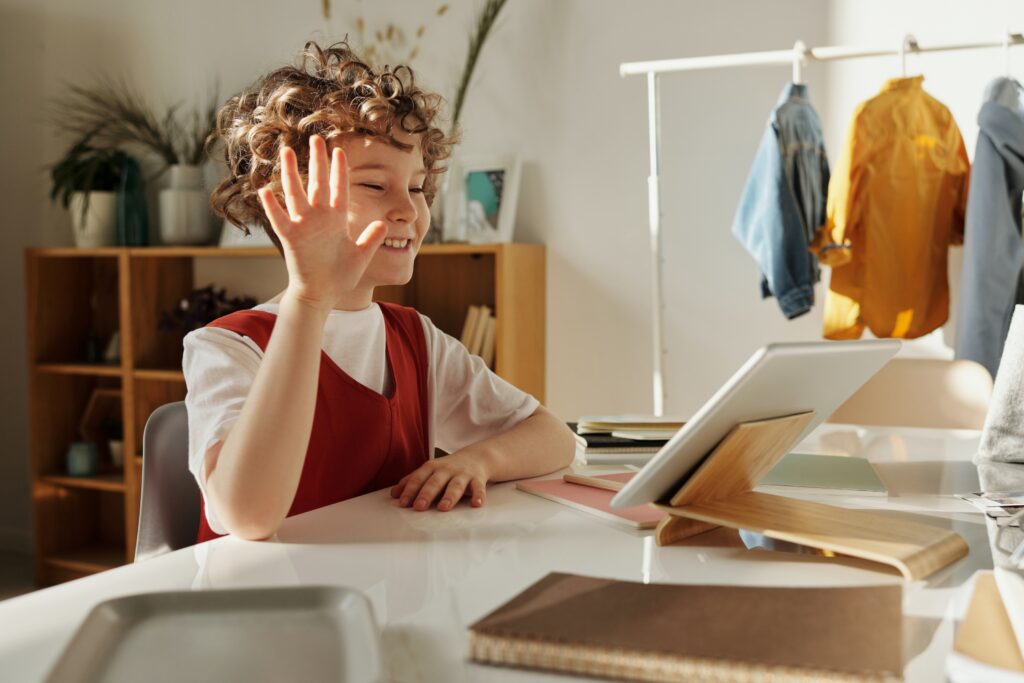 A young boy smiles and waves at the camera while seated at a desk, creating a cheerful and engaging atmosphere.